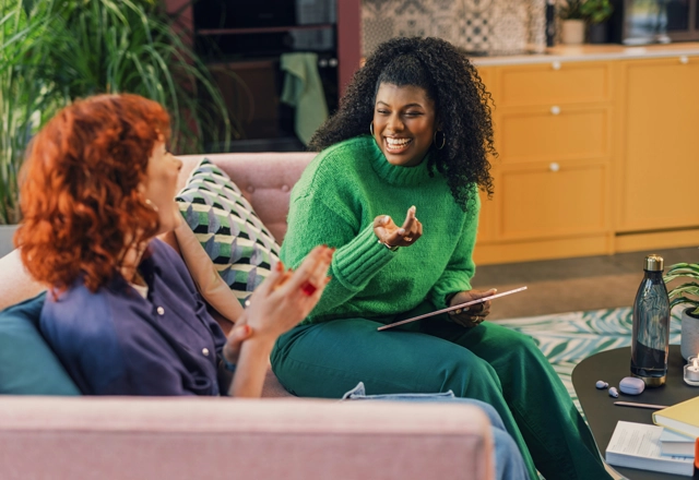 Two women sitting on pink sofa and laughing and one of them is holding a tablet.
