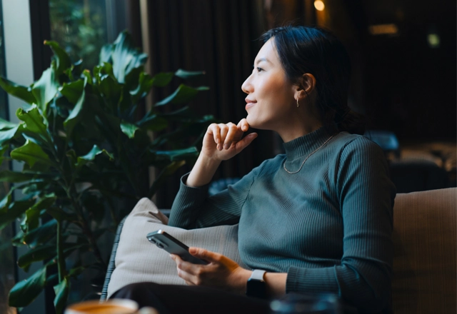 Woman sitting and looking out a window.  Plants are behind her and she has a phone in hand.